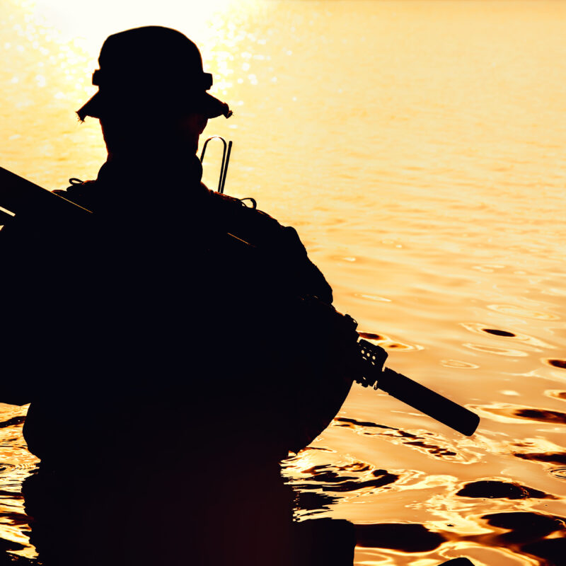 Silhouette of special forces with rifle in action during river raid in the jungle waist deep in the water. Front view, half length
